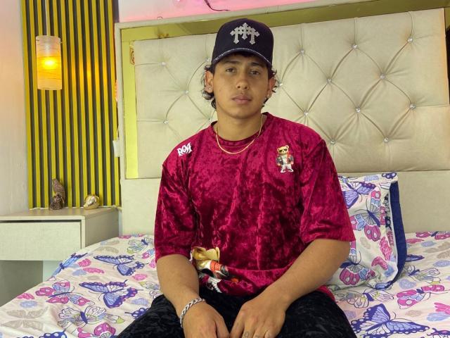 AxelMonroe sits on a bed with a tufted, beige headboard. He wears a dark blue baseball cap with a white emblem, a red velvet shirt with small logos, and black pants. The bed has a white sheet with blue and purple butterfly patterns and a matching pillow. A white nightstand with a yellow-striped wall is on the left. Warm, yellow light from a wall sconce is visible. AxelMonroe has short, curly hair and a neutral expression.