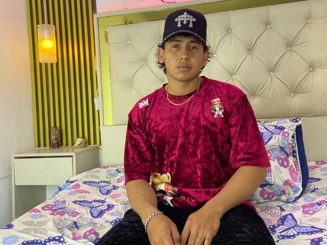AxelMonroe with curly brown hair, wearing a black cap, red velvet shirt, black pants, and a silver bracelet, sitting on a butterfly-patterned bed. He has a neutral expression.