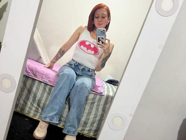 Valeriaaf with red hair, white Batman tank top, blue jeans, and pink sneakers, sitting on a bed, holding a phone, showing tattoos on arms, mirror selfie.