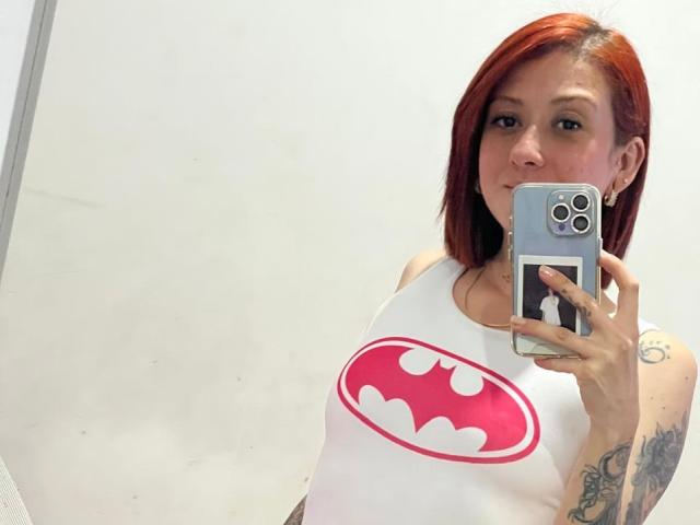 Valeriaaf with red hair, holding phone, wearing white tank top with large pink Batman logo, showing her tits, tattoo on right arm, in mirror selfie.
