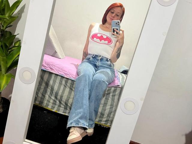 Valeriaaf with red hair, white Batman tee, blue jeans, and white sneakers, sitting on a bed, taking a mirror selfie, showing a tattoo on her left arm.