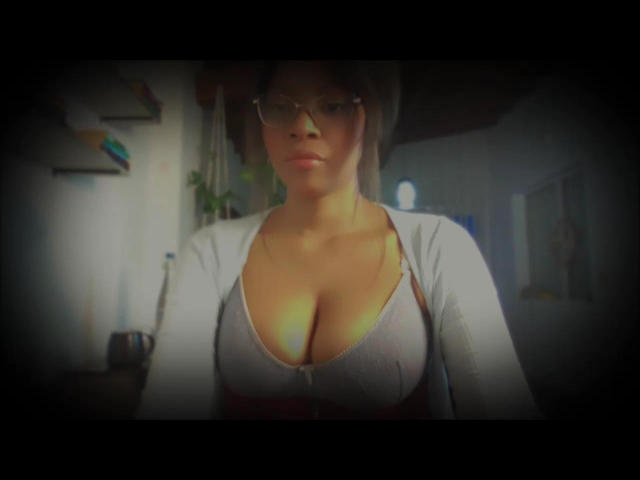 Adouxrable with big boobs in a white cardigan and glasses, looking down, has brown hair and a serious face, standing in a dimly lit room.