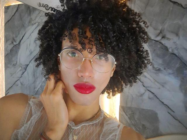 CamilleCedrik has curly black hair and wears gold-rimmed glasses. She has fair skin and red lipstick. Her right hand touches her cheek. She wears a sheer white lace top. The marble background has gray and white veins. The lighting is bright, highlighting her face. The image is a close-up, focusing on her face and upper torso. Her curly hair is textured, and her glasses have a slight reflection. She has a nose ring on her right nostril.