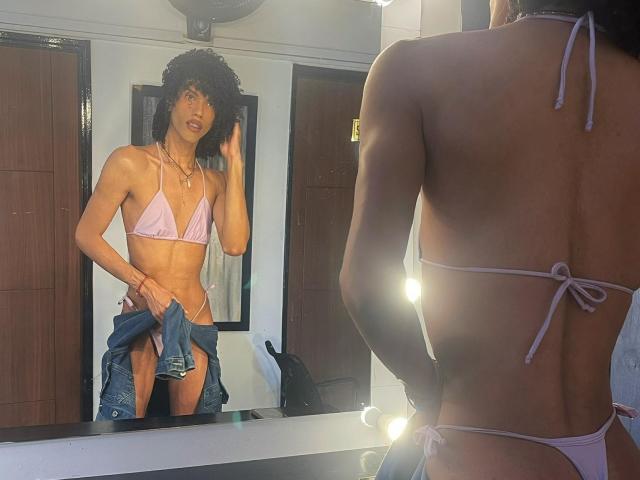 CamilleCedrik stands in front of a mirror, wearing a pink string bikini that highlights their slim, toned physique. They have curly black hair and a light brown skin tone. CamilleCedrik holds a blue denim jacket over their right hip with their left hand, looking into the mirror with a slightly parted lip. The mirror reflects their front, showing the bikini's triangular top and side ties. Bright bulbs are on the mirror's right side, casting light on CamilleCedrik's back. The room has a wooden door in the background.