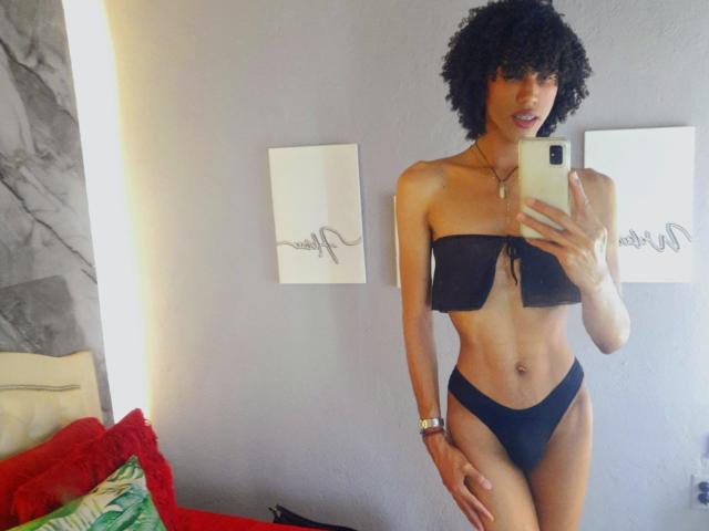 CamilleCedrik, met krullend zwart haar, draagt een zwart, strapless bikini topje en een bijpassende broek, en houdt een telefoon vast. Ze laat een slank, getraind lichaam zien met kleine borsten en een platte bil..