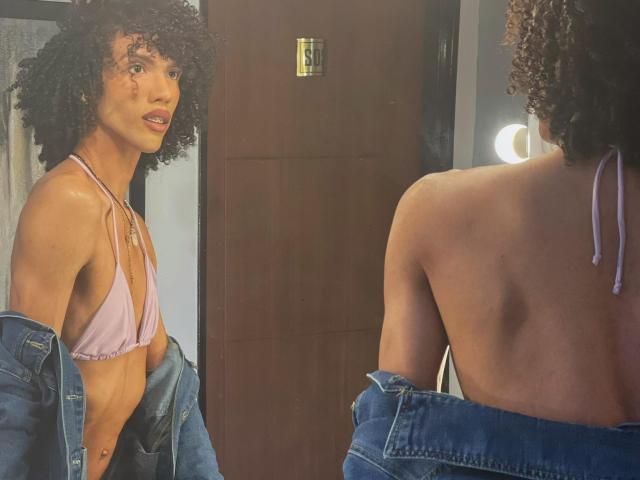 A photograph of CamilleCedrik with curly hair and a light brown skin tone. She wears a pink bikini top, a blue denim jacket, and has a slim, toned physique. She's facing a mirror, showing her back to the camera. The mirror reflects her front, highlighting her facial features and a small scar on her cheek. A round mirror light is visible in the reflection. The image has a warm, natural light.