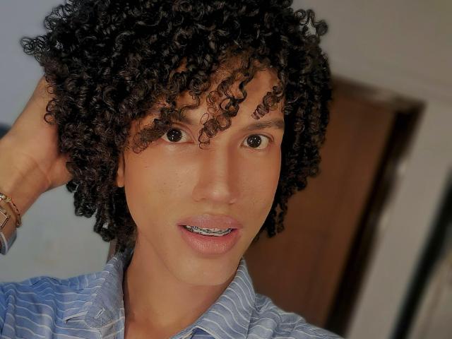 Close-up of CamilleCedrik with curly black hair, medium brown skin, and brown eyes. CamilleCedrik is looking slightly to the right, mouth open with visible teeth and a slight smile. Wearing a blue striped shirt, left hand touches hair. Natural light highlights skin texture. Brown door in the blurred background. Minimalist composition, focus on facial features. Slightly messy hair, soft shadows. Casual, everyday look. Simple bracelet on left wrist. Image has a realistic, candid feel.