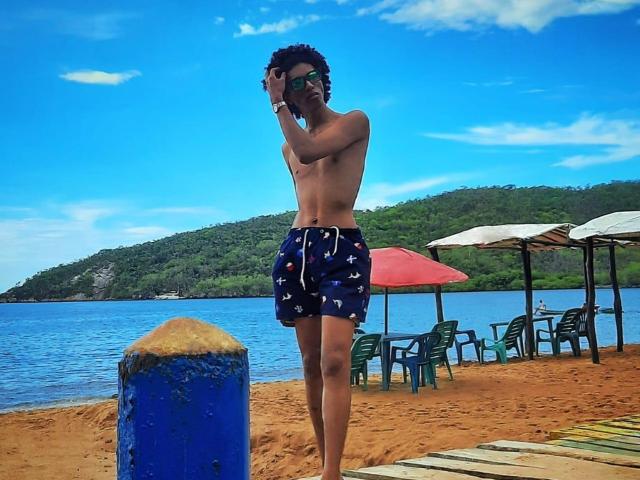 CamilleCedrik stands on a sandy beach, shirtless, wearing dark blue swim shorts with a white pattern. He has curly hair and is adjusting his sunglasses with his right hand. His skin is medium brown. In the background, green hills and a blue sky with scattered clouds are visible. There's a blue post in the foreground and a red umbrella with green chairs under it. The beach has a wooden walkway on the right. The image is vibrant with strong contrasts between the blue sky, green hills, and sandy beach.