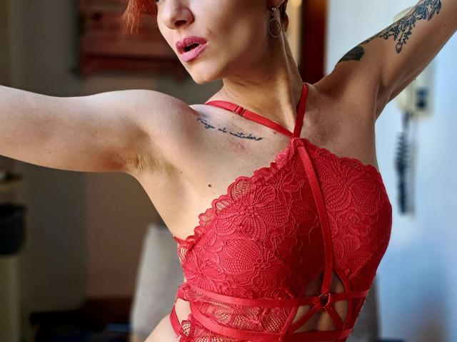 DreamityCamy trägt ein rotes Spitzen-Bralette, das ihre Brüste zeigt, mit einem Tattoo an der rechten Schulter, einem erhobenen Arm, roten Haaren, Lipgloss und Ohrringen. Weiches, natürliches Licht, leicht geöffneter Mund..
