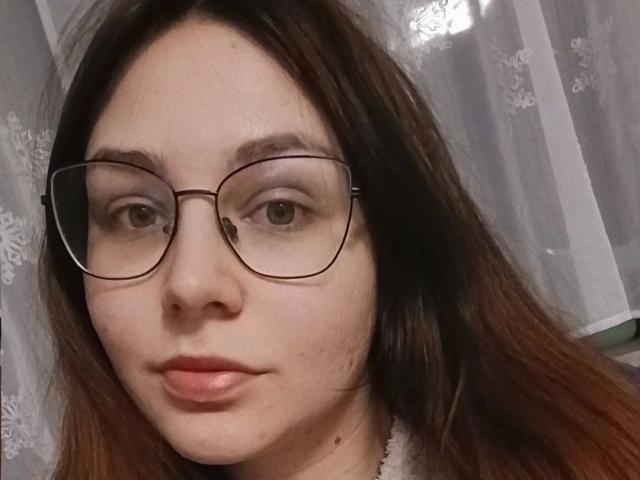 Close-up of MiaEngel-hot with long brown hair, wearing glasses with thin, brown metal frames. Her skin is fair with a few small moles on her chin. She has light brown eyes and neutral, slightly pink lips. The lace curtain in the background is sheer and white, adding texture. The photograph has a soft, natural light that highlights her facial features. The focus is on her face, with a slight tilt to the right. Her expression is calm and slightly serious.