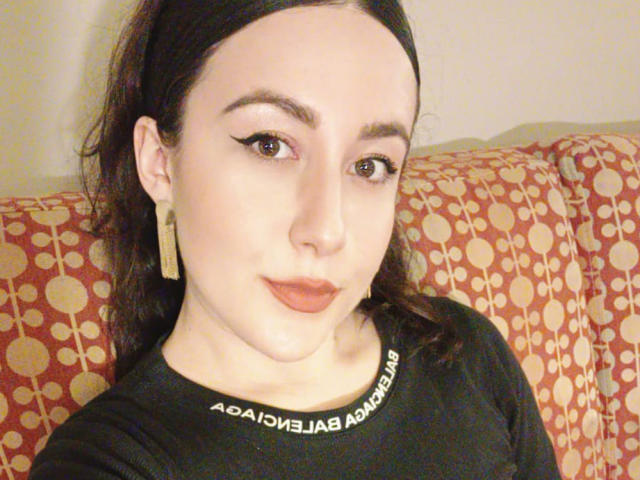MabyLegrand has pale skin, dark brown hair, winged eyeliner, red lipstick, and gold earrings. She's wearing a black shirt with white text. Her face is slightly chubby.