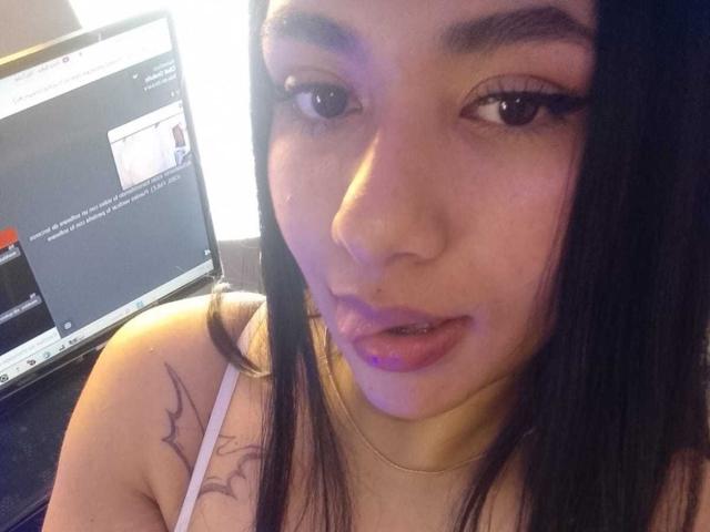 KaileeRoss has a tattoo on her shoulder, dark eyeliner, and purple lipstick, with her tongue slightly out, looking at a laptop screen in a dimly lit room.