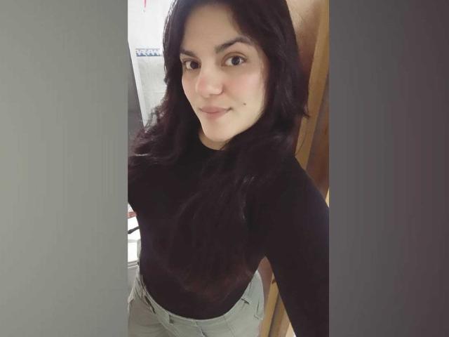 PaulinaCox with dark wavy hair, wearing a black long-sleeve top and light grey high-waisted pants, smiling subtly at the camera, showing a cute small mole on her right cheek.