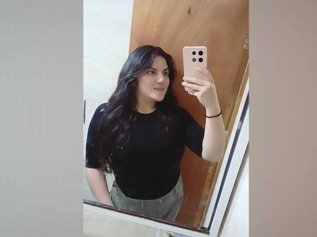 PaulinaCox with long black hair, wearing a black top and gray pants, holding a pink phone, taking a mirror selfie, showing a small smile.