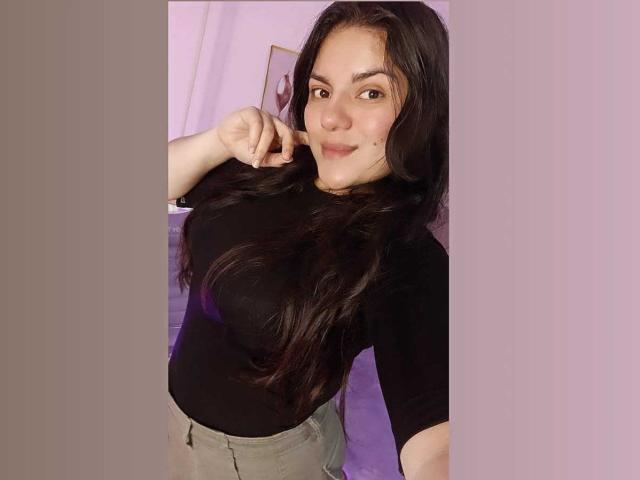 PaulinaCox with long black hair, wearing a black top, pointing at her nose, grey pants, and a cute smile, showing her big tits. Her brown eyes are focused.