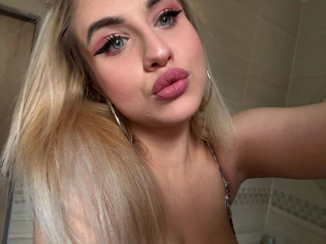 LolaLove-hot with pink lipstick, blonde hair, and hoop earrings, pouting with her plump, shiny, nude-colored tits barely visible in a strap top, in a bathroom selfie.