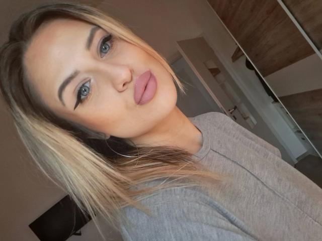 LolaLove-hot with blonde hair, blue eyes, and pink lips, wearing a gray shirt, pouting, in a selfie. Her tits are not visible.