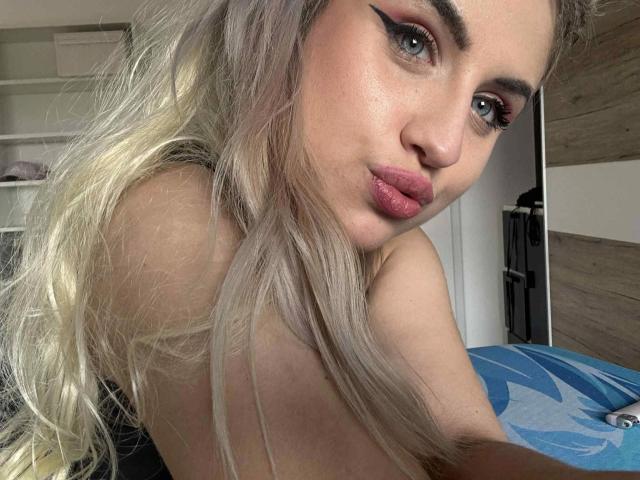 LolaLove-hot with blonde hair, blue eyes, and pink lips, showing off her bare shoulders and pouty face, wearing bold winged eyeliner. Her tits are partially visible.