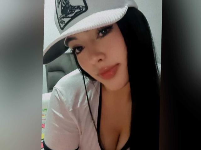 CamillaRiley with long black hair, wearing a white cap and low-cut shirt, showing her boobs and a slight smile. She has light skin and full pink lips.