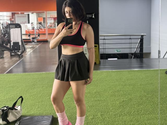 AnastasiaEvansX in a gym, mirror selfie, wearing a black sports bra with pink trim, black pleated skirt, pink socks, holding phone, showing toned legs, small tattoos on left arm.