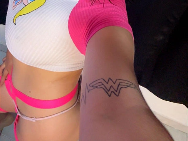 AnastasiaEvansX's arm with a bold "WW" tattoo, wearing a pink and white striped top, pink thong, showing her tan skin and slim waist. Her pink thong is visible.