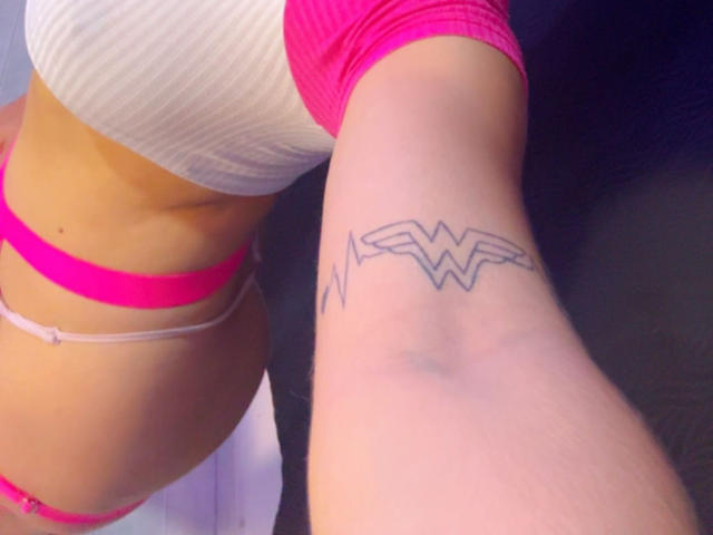 AnastasiaEvansX's arm with a black "WW" tattoo, wearing pink and white lingerie, showing her tan skin and small pink thong, with her pink bra-covered tits visible.