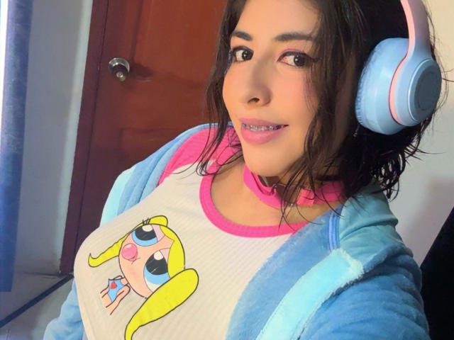 AnastasiaEvansX with pink lipstick, blue headphones, wavy brown hair, wearing a pink and white shirt with a cute blonde cartoon character, and a blue hoodie. She's smiling.