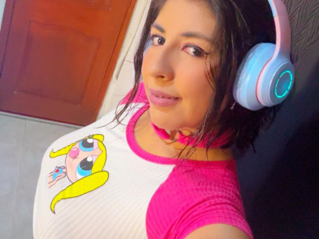AnastasiaEvansX with shoulder-length dark brown hair, wearing pink headphones with a blue accent, a pink choker, and a white raglan shirt with a pink hem and a cartoon yellow bird on the chest. She has light brown skin, pink lipstick, and a slight smile. The image has a bright, colorful palette with a focus on pink and yellow. The background includes a wooden door and a gray floor. The composition is a close-up selfie.