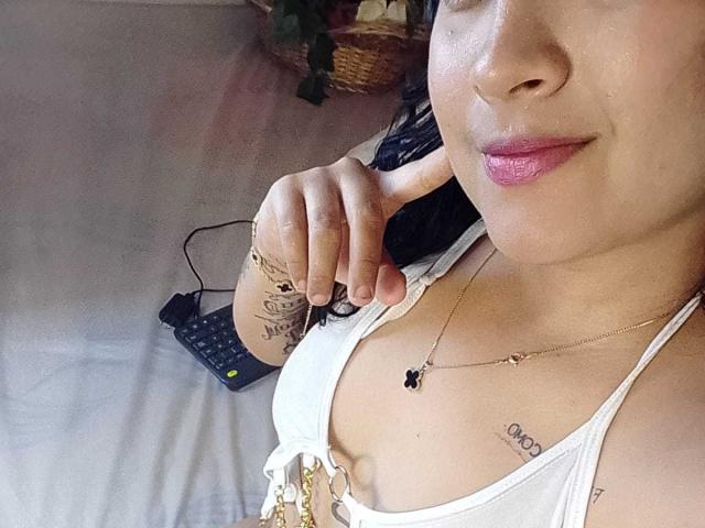 DanniWest has pink lipstick, white top, showing tits, tattoos on her wrist and chest, finger pointing, black hair, gold necklace, remote control on bed, slightly smiling.