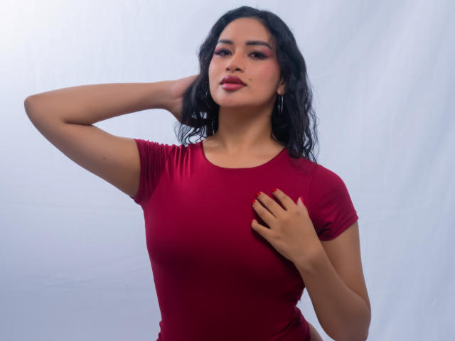 AdalinaVera with dark wavy hair, wearing a tight red shirt, has one hand on her head and the other on her chest, showing her small tits and red nails.