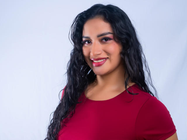 AdalinaVera with wavy black hair, red shirt, and pink eye makeup, smiling with red lipstick, showing her tits in a portrait.
