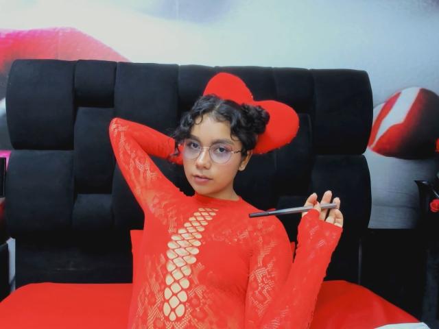 JuliaBennett with curly black hair, glasses, and red lace dress with a heart-shaped hat, holding a pen, sitting on a black tufted sofa, looking relaxed.