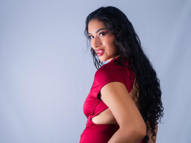 AdalinaVera with long black curly hair, red dress, showing sideboob, looking over shoulder, smiling, red lipstick, light skin, standing against light blue background.