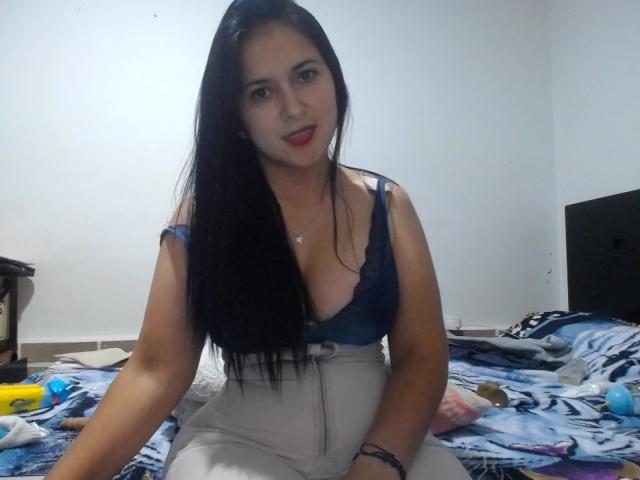 PrinceHermosa, with long black hair, wearing a blue lace bra and white pants, sitting on a bed with blue patterned sheets, showing her tits and smiling slightly.