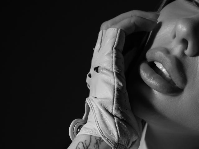 Close-up black and white photo of SensualMila's face. She's biting her lower lip, showing her teeth, with a finger from her right hand gripping the strap of a white leather glove that's partially torn. Her lips are full, slightly parted, and her skin has a smooth texture. A tattoo is visible on her left forearm. The image is sharply focused on her face and glove, with a dark, out-of-focus background. The composition emphasizes the tension between her lips and the strap.