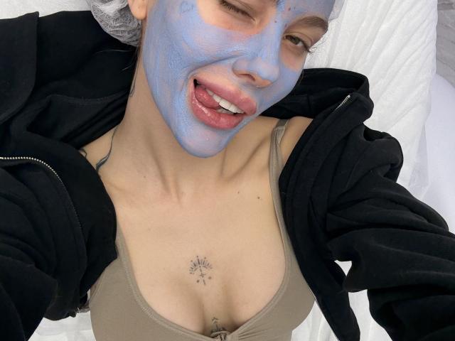 SensualMila heeft een blauw gezichtsmasker, knipoogt met haar tong uit, een zwarte hoodie, een bruine bh, toont kleine borsten met een sterretje tatoeage erboven, en een zwarte haarcap..