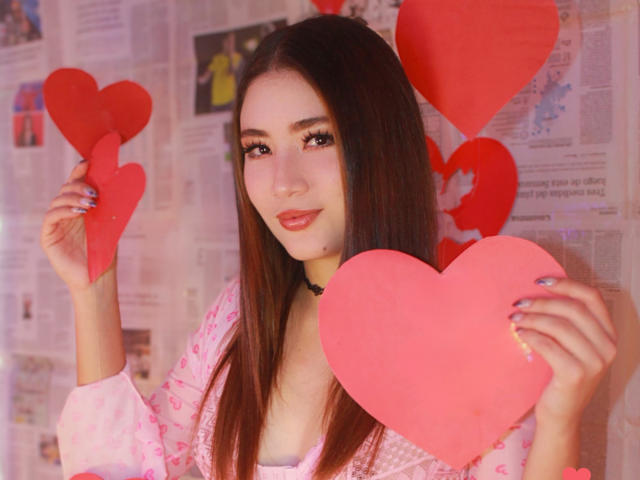StarDustt with long brown hair, pink heart-shaped papers in hands, wearing pink patterned shirt, black choker, smiling, holding hearts in front of face. Red hearts in background.