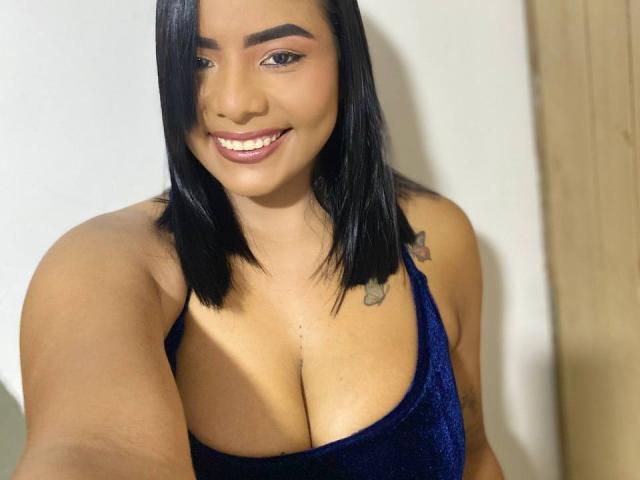 SilverLuna with medium brown skin, black shoulder-length hair, smiling, wearing a blue velvet dress, showing her big tits, tattoo on right shoulder, in a selfie.