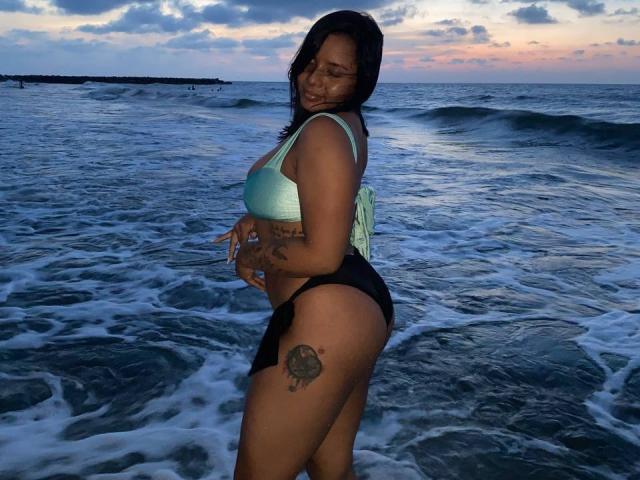 SilverLuna with tan skin, black hair, green bikini top, black bikini bottom, tattoos on thigh and side, standing in ocean, waves, sunset, smiling, looking over shoulder.