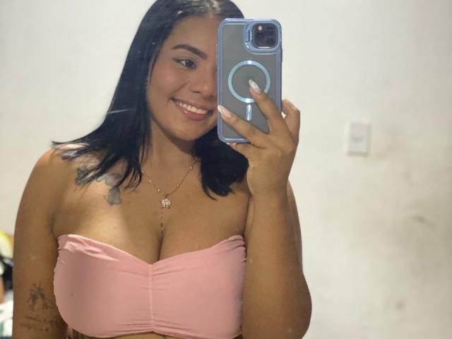 SilverLuna, con la pelle marrone media e i capelli neri, sorride e sta scattando un selfie con un telefono blu. Indossa un top rosa senza spalline, che mostra il seno e un tatuaggio sul braccio..