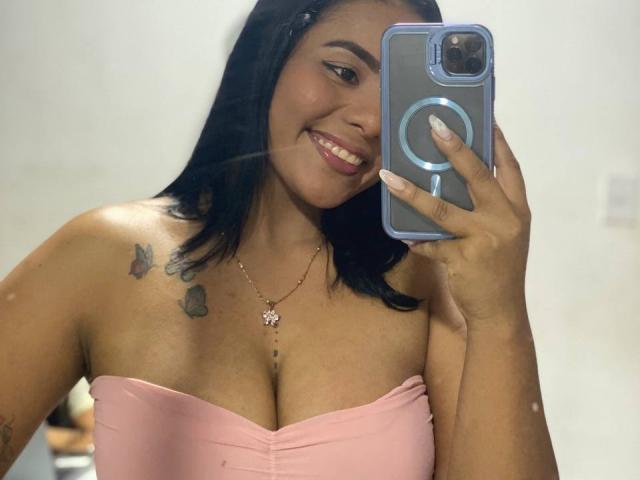 SilverLuna with black hair, pink strapless top, smiling, holding phone, visible tits, butterfly tattoos on right shoulder, necklace with butterfly pendant, taking selfie.