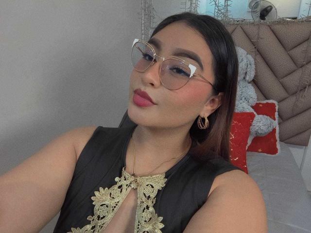 HelueJamil con gafas, lápiz labial rosa, vestido negro con bordados dorados, aretes de aro dorados, sentada, sonriendo ligeramente, cabello castaño, piel clara, dormitorio, selfie..