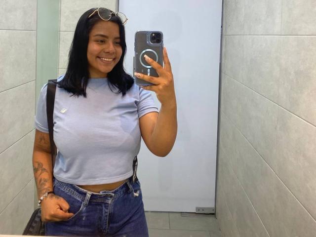 SilverLuna, con i capelli neri, sorridente, indossa una maglietta azzurra, jeans a vita alta, tiene in mano un telefono, ha tatuaggi visibili sul braccio, occhiali sulla testa, è in piedi in un bagno..