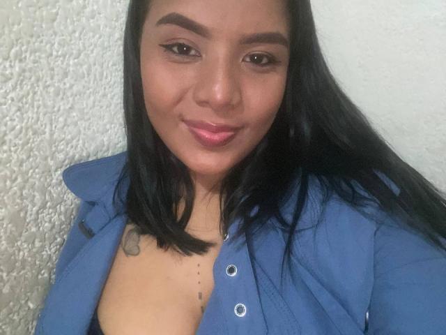 SilverLuna, com cabelo castanho escuro, batom rosa e camisa azul, mostrando seios pequenos e uma tatuagem no ombro esquerdo, sorrindo para a câmera. Parede branca texturizada ao fundo..