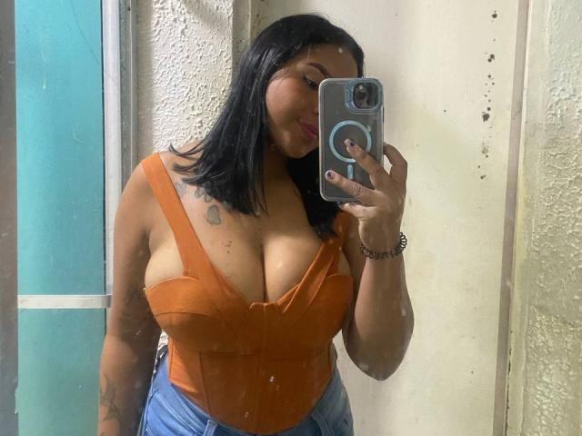 SilverLuna with black shoulder-length hair, orange deep-cut top showing big boobs, blue jeans, holding phone in mirror selfie, pink lipstick, tattoo on left shoulder, smiling.
