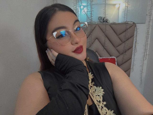 HelueJamil with red lipstick, black lace dress, gold embroidery, and glasses, resting her chin on her hand, showing off her tits, with a confident, sexy look.