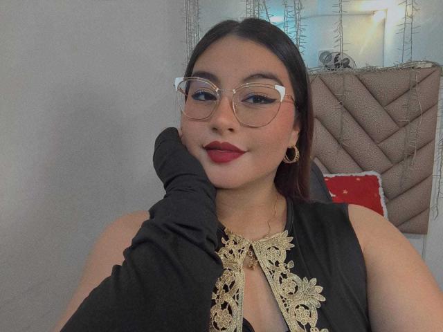 HelueJamil with glasses, black dress, gold lace, and red lipstick, black glove on cheek, small earrings, smiling, brown hair, medium tits, fair skin, indoors.