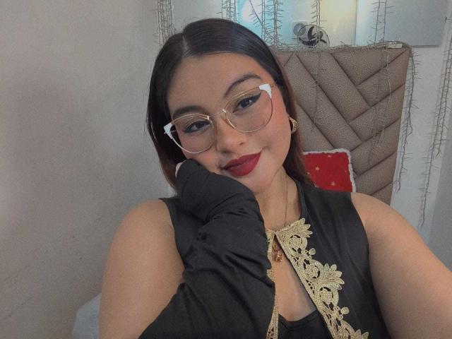 HelueJamil with red lipstick, glasses, black sleeveless top with gold embroidery, black glove, and dark hair, smiling at the camera. Her tits are partially visible under the top.