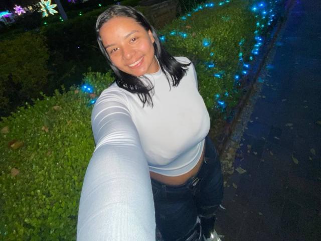 SilverLuna, with medium tits, wearing a white long-sleeve top, dark jeans, and black boots, takes a selfie at night, smiling, with blue lights in the background.