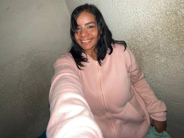 SilverLuna with dark wavy hair, smiling, wearing a pink hoodie, extending her arm for a selfie, showing her tits in the foreground, against a textured wall.